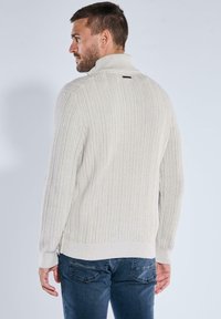 Hellgrauer gerippter Pullover mit hohem Kragen und strukturierter Stoffoberfläche, versehen mit einem kleinen schwarzen Akzent am Ausschnitt. Trägt er mit dunklen Jeans.