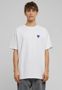 Hvid bomulds t-shirt med et blåt hjerteemblem på venstre bryst. Blød tekstur, afslappet pasform, korte ærmer og rund halsudskæring.
