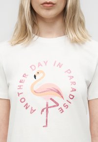 Ξανθιά γυναίκα που φοράει λευκό μπλουζάκι με γραφικό ροζ φλαμίνγκο και το κείμενο «Άλλη μια μέρα στον παράδεισο» με ροζ γράμματα γύρω από αυτό.