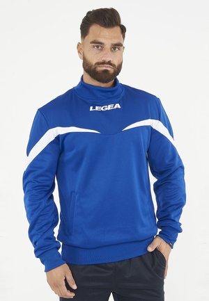 Muž s vousy, který nosí modrý sportovní vršek s bílými pruhy a logem "LEGEA", stojí s jednou rukou v kapse.