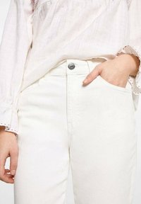 Bijele traperice od denima slim kroja, s metalnim gumbom za zatvaranje i prednjim džepom. Tkanina ima glatku teksturu.