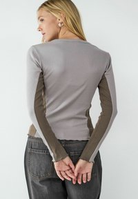 Langsleeve top met een geribbelde textuur in lichtgrijs, voorzien van donkergrijze zijpanelen en onafgewerkte zoomdetails; gecombineerd met donkere jeans.