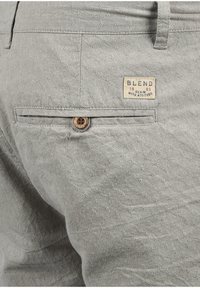 Ljust grå texturerade byxor med detalj av en knappad bakficka och en liten rektangulär lapp med texten "Blend Denim with Attitude".