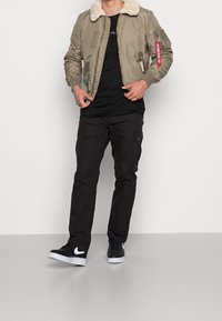 Giacca bomber khaki con colletto in pelliccia sintetica, polsini a costine e molteplici tasche con zip, abbinata a una t-shirt nera e pantaloni cargo.