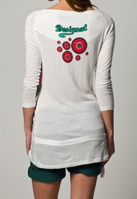 Vits långärmad topp med rundad sidsöm, prydd med grön "Desigual" logotyp och färgglada cirkulära mönster på baksidan. Mjuk bomullstextur.