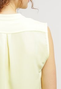 Sleeveless light yellow blouse with a smooth texture, classic collar, and a back pleat detail. Satin-like fabric with no visible embellishments.