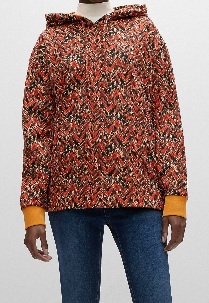 Sudadera con capucha que presenta un patrón en zigzag en rojo, negro y crema, con puños naranjas y un corte relajado. Fabricada en tejido suave.