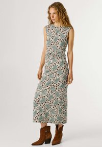 Ženska z dolgimi kodrastimi lasmi, oblečena v brezrokavno cvetlično midi obleko in rjave semišne gležnjarje, stoji na belem ozadju.