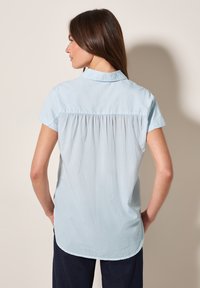 Chemise bleu clair à manches courtes avec un col, présentant des rayures verticales discrètes et un détail froncé dans le dos.