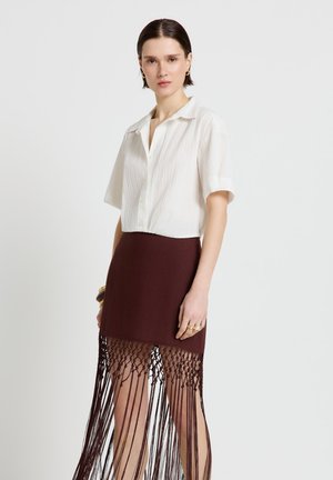 Frau trägt ein weißes, kurzärmeliges Hemd mit Knöpfen und einen braunen Rock mit langen Fransen, steht vor einem schlichten Hintergrund.