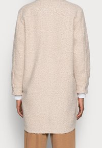 Persona che indossa un lungo cappotto di lana beige, testurizzato, sopra delle maniche bianche e pantaloni marroni, mostrata di spalle su uno sfondo semplice.