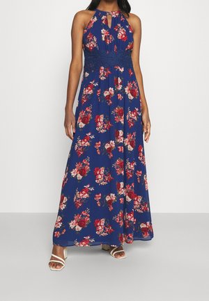 Kobieta ubrana w niebieską, bezrękawnikową suknię maxi z czerwono-białym kwiatowym wzorem oraz białe sandały na obcasie z paskami, stojąca na jednolitym tle.