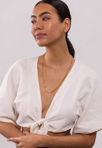 Eine goldene Halskette mit kreisförmigen Gliedern, getragen über einer weißen, strukturierten Bluse mit Bindeband. Die Bluse hat kurze Ärmel und ein tiefes V-Ausschnittdesign.