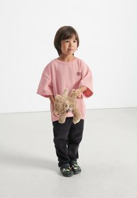 Barn, der bærer en løs lyserød bomulds T-shirt med et logo, sorte corduroy bukser og sorte dekorative klip-klapper, mens det holder en tan-farvet stuffed bjørn.