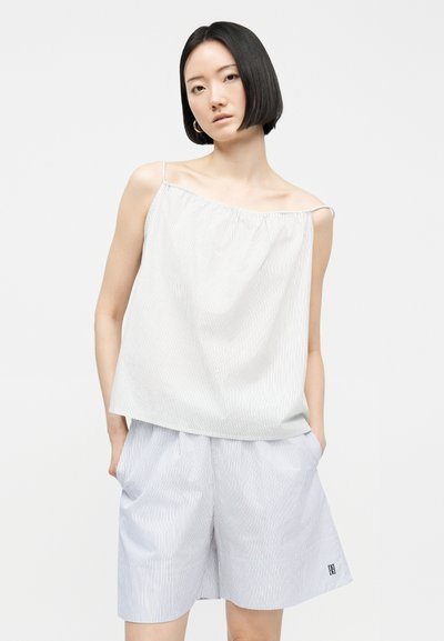 Γυναίκα με κοντά μαύρα μαλλιά, ντυμένη με άνετο λευκό αμάνικο τοπ με ρίγες και σορτς, με τα χέρια στις τσέπες, μπροστά σε απλό φόντο.