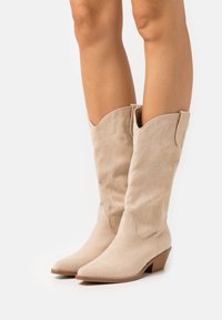 Beige suède cowboylaarzen met een spitse neus en een lage stapelhak. Voorzien van decoratieve stiksels langs de bovenste schacht en zijpanelen.