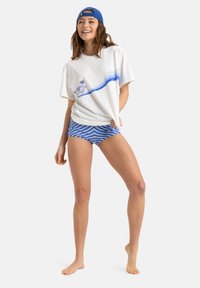 Vit t-shirt med blå grafik av palmer och vågor, i kombination med blå och vita randiga shorts. En blå keps fulländar outfiten.