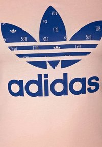 Camiseta rosa con un gran logo de Adidas en azul, adornado con un diseño de hojas y patrones geométricos, fabricada en un tejido suave y liso.