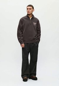 Άνδρας φορεί σκοτεινό γκρίζο φούτερ με φερμουάρ έως το τέταρτο και λογότυπο, μαύρο παντελόνι και μαύρα παπούτσια, στέκεται με τα χέρια στις τσέπες σε υπόβαθρο χωρίς σχέδιο.