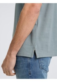 T-shirt gris clair à manches courtes avec une finition texturée, des fentes sur les côtés et une coupe décontractée, associé à un jean en denim bleu.