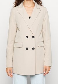 Beige dubbelknäppt kavaj med svarta knappar, slagkragar, ficklock, bärs över ljusblå jeans på en person med lockigt hår.