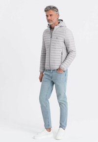 Polstret grå jakke med hætte, der har horisontale syninger, lynlåslommer og elastiske manchetter. Parret med lyseblå jeans og hvide sneakers.