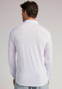 Camisa polo blanca de manga larga con una textura suave, un cuello clásico y detalles en los puños con botones, mostrada desde la parte trasera.