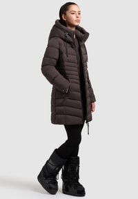Dark brown puffer coat with a hood, featuring zippered pockets, a slightly tapered design, and textured quilting, paired with black winter boots.