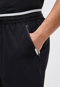 Ranka įkišta į užtrauktuku užsegamą juodos spalvos sportinių kelnių šoninę kišenę, kurios juosmuo elastingas, o šonuose yra baltos spalvos juostelės detalės.