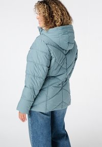 Lichtblauwe gewatteerde jas met een capuchon, gekenmerkt door diagonale gestikte patronen en een ritszak op de borst. Draagt een paar wijde denim jeans.