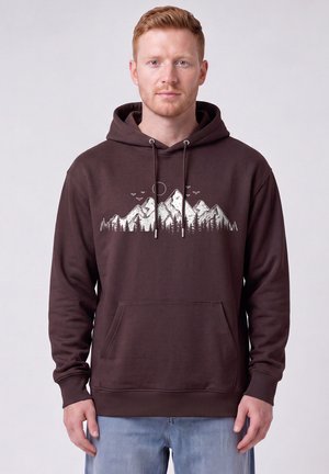 Man met kort rood haar, gekleed in een donkerbruine hoodie met berg-, boom-, zon- en vogelontwerp op de borst, staand tegen een effen achtergrond.