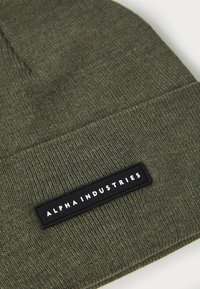 Olivově zelená pletená čepice s texturovaným povrchem, která má černou obdélníkovou nášivku s nápisem "ALPHA INDUSTRIES" bílým písmem.