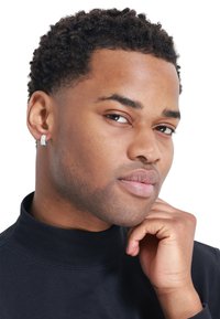 Silver hoop earring with small gemstones, worn by a person in a black turtleneck. Smooth skin texture and curly dark hair visible.