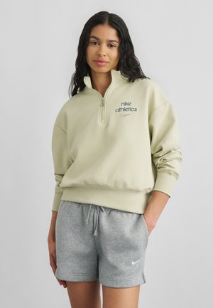 Jeune femme portant un sweat-shirt Nike Athletics beige clair à fermeture éclair quart de zip et un short gris Nike à cordon, debout avec une main dans la poche.