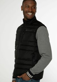 Czarna pikowana kamizelka z zamek błyskawicznym, z szarą koszulą z długim rękawem pod spodem, w paskowane mankiety i małym logo na piersi.