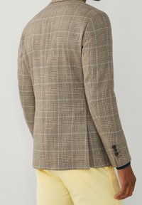 Brązowo-szary blazer w kratę, teksturowany materiał, dopasowany krój, z wyłogami i detalami z trzema guzikami na mankietach, w zestawieniu z jasnobeżowymi spodniami.