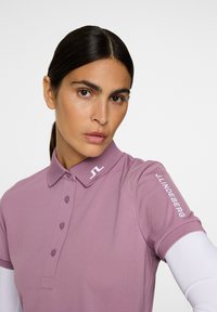 Lila polo-skjorta med klassisk krage, knappstängning och vit JL-logga på ärmen. Slät textur och figursydd design.