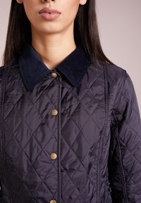 Navy gewatteerde jas met een corduroy kraag, gouden drukknopen en een ruitvormig stikpatroon; gladde textuur, getailleerd ontwerp.