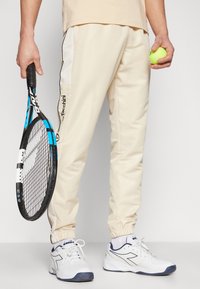 Gaišbeidzs sporta bikses ar elastīgām aprocēm, kontrastējošām melnām sānu joslām un logotipa detaļām. Tur rokā tenisa raketi un zaļu tenisa bumbu.