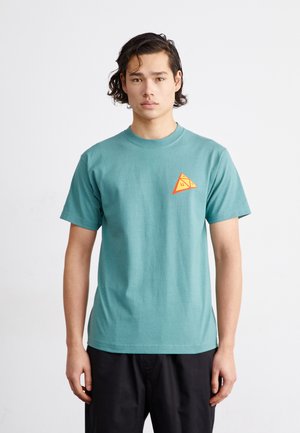 Camiseta de algodón verde azulado con cuello redondo, que presenta un pequeño logotipo en forma de triángulo naranja en la parte izquierda del pecho. Mangas cortas.