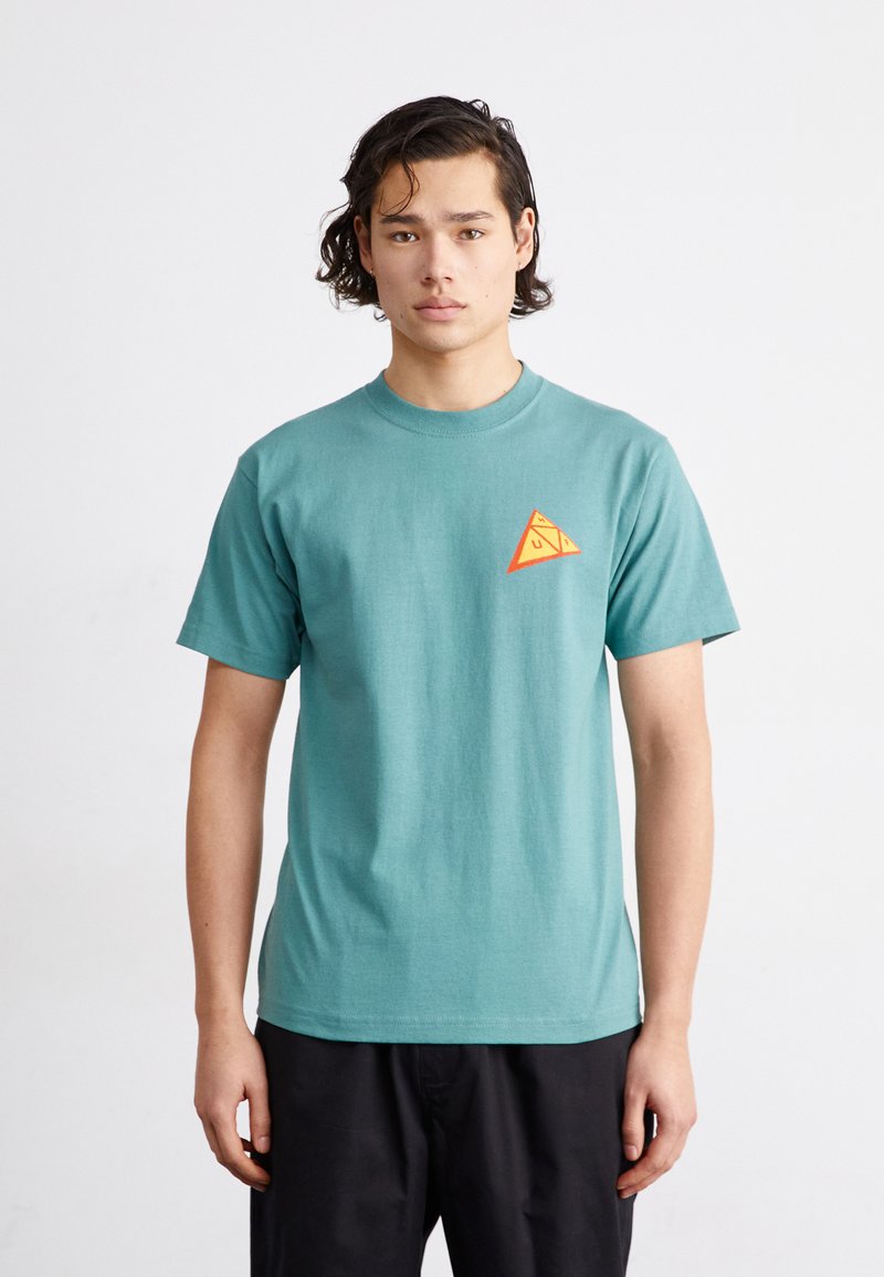 Camiseta de algodón verde azulado con cuello redondo, que presenta un pequeño logotipo en forma de triángulo naranja en la parte izquierda del pecho. Mangas cortas.