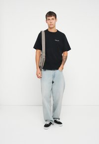 Sort bomuld t-shirt med hvid tekst, lyseblå løstsiddende denim jeans, sorte sneakers og en grå crossbody taske. Ensfarvet baggrund.