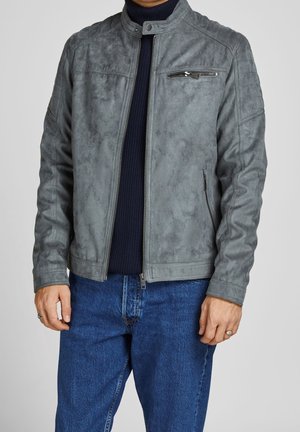 Man draagt een grijs suède jack met ritssluiting over een zwarte coltrui en blauwe jeans, staand met ontspannen handen langs het lichaam.