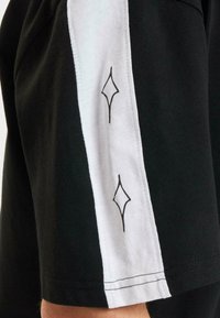 Juodas medvilnės marškinėlis su baltu šoniniu brūkšniu, turinčiu deimanto raštą ir kontrastingą siuvimą. Marškinėliai pasižymi laisvu siluetu.