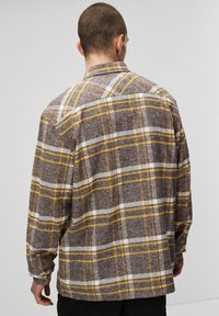 Brązowa flanelowa koszula w żółto-białą kratę, z długimi rękawami i luźnym krojem, z tylnym karczkiem i guzikami przy mankietach.