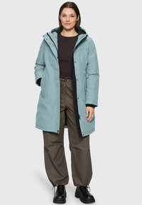 Vrouw in een lichtblauwe gewatteerde jas met capuchon, draagt een zwart shirt, bruine broek met trekkoord en zwarte laarzen, staande tegen een simpele achtergrond.