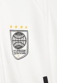 Hvidt stof med broderet sort globe inden i et skjold, fem gule stjerner over, og tekst der lyder "STORM DESIGN STUDIO WORLDWIDE CREATIONS."