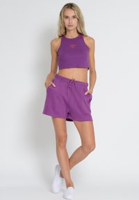 Purple cropped tank top with ribbed texture and logo, paired with matching purple drawstring shorts featuring pockets. White slip-on shoes.