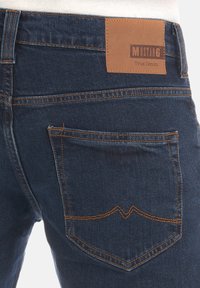 Jeans de mezclilla azul oscuro con costuras naranjas, que cuentan con una etiqueta de cuero en la cintura y un bolsillo trasero con una costura de forma distintiva.