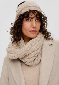 Beige gebreide sjaal met een gevlochten structuur, gecombineerd met een bijpassende hoofdband. Draagt over een lichtbeige trui en een getailleerde jas.